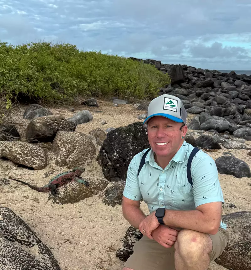 AdventureSmith traveler kneeling down next to a Christmas iguana on the rocky beach on a bright sunny day