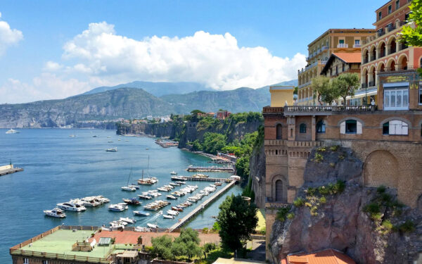 Coastal town of Amalfi with boats docked in a marina, cliffs, and buildings overlooking a blue sea under a partly cloudy sky.