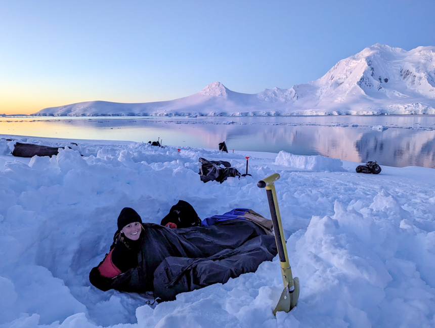 Women in sleeping bag snow camping off the coast of Antarctica with the sunsetting on a calm day