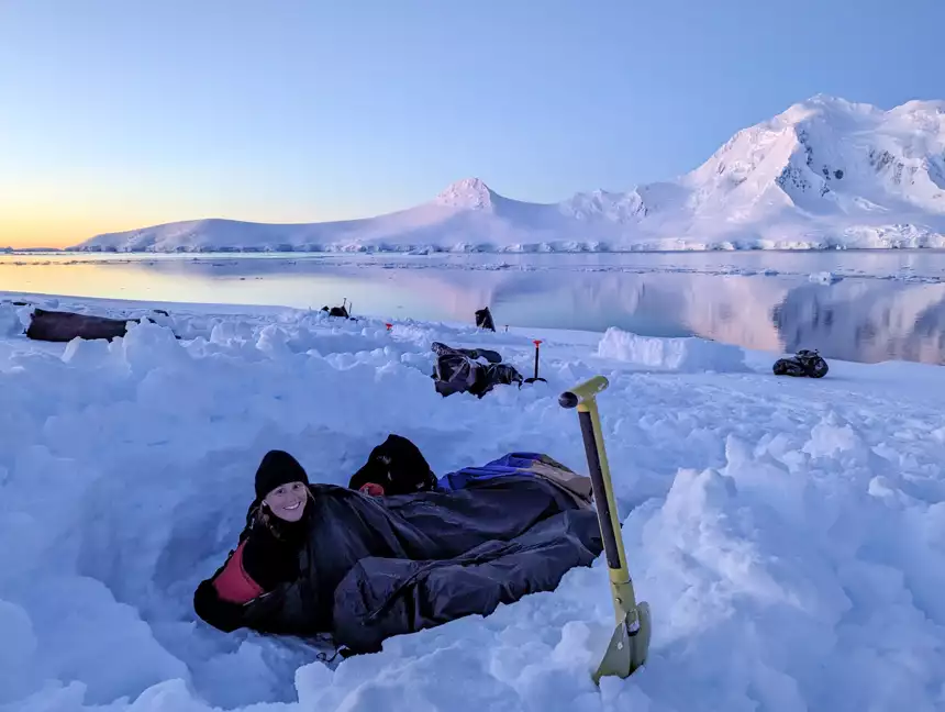 Women in sleeping bag snow camping off the coast of Antarctica with the sunsetting on a calm day