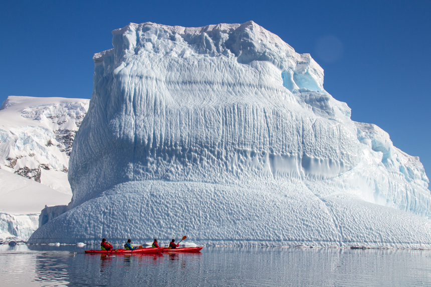 Kayakers paddling through the calm waterways of Antarctica next to massive iceberg on a sunny day