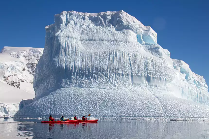 Kayakers paddling through the calm waterways of Antarctica next to massive iceberg on a sunny day
