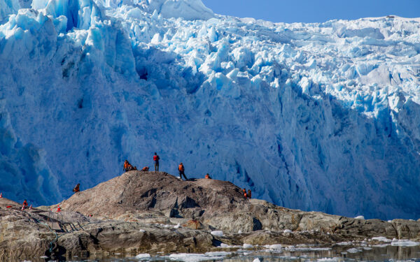 A group of travelers on a Chile adventure trip stand on rocks in front of a massive blue glacier taking up the whole image