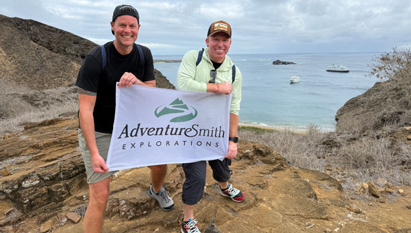 2 men in warm weather attire hold a white & green flag on a clifftop overlooking a small cove with 2 small ships in the Galapagos.