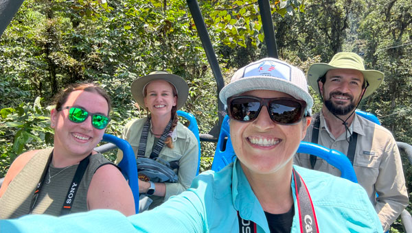 Woman in teal shirt holds camera for a selfie with 3 other travelers riding among the jungle canopy during an Ecuador vacation.