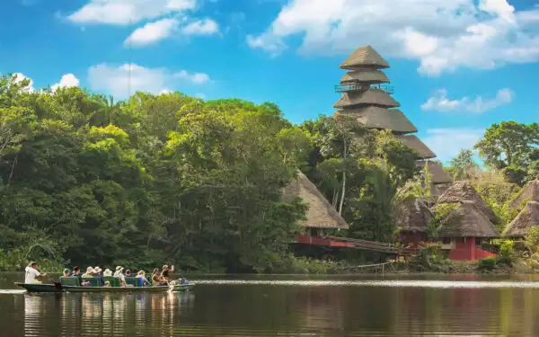 A canoe filled with an adventure tour group paddles towards a multi-level Amazon lodge with thatched roof