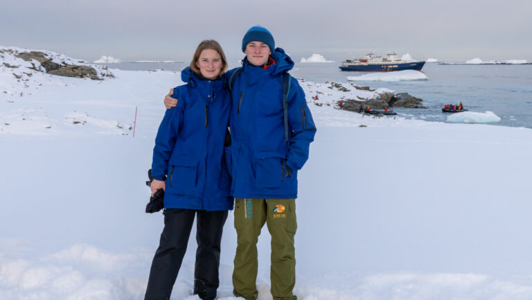 Young man & woman in blue jackets stand on snowfield by calm sea with a blue & white small ship in the distance in Antarctica.