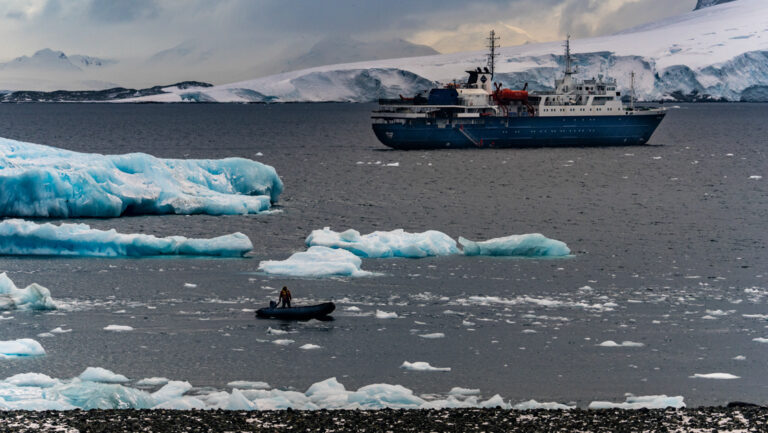 Small ship with blue hull & white upper decks sits in calm sea among blue icebergs as Zodiac cruises nearby in Antarctica.