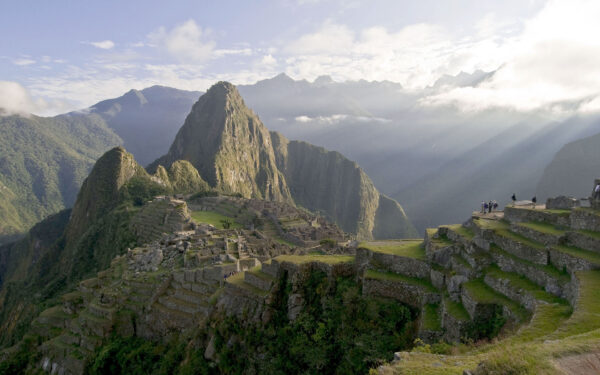 Sunbeams shine down on Machu Picchu and Huayna Picchu with a guided tour group seen standing on rock walls above the citadel
