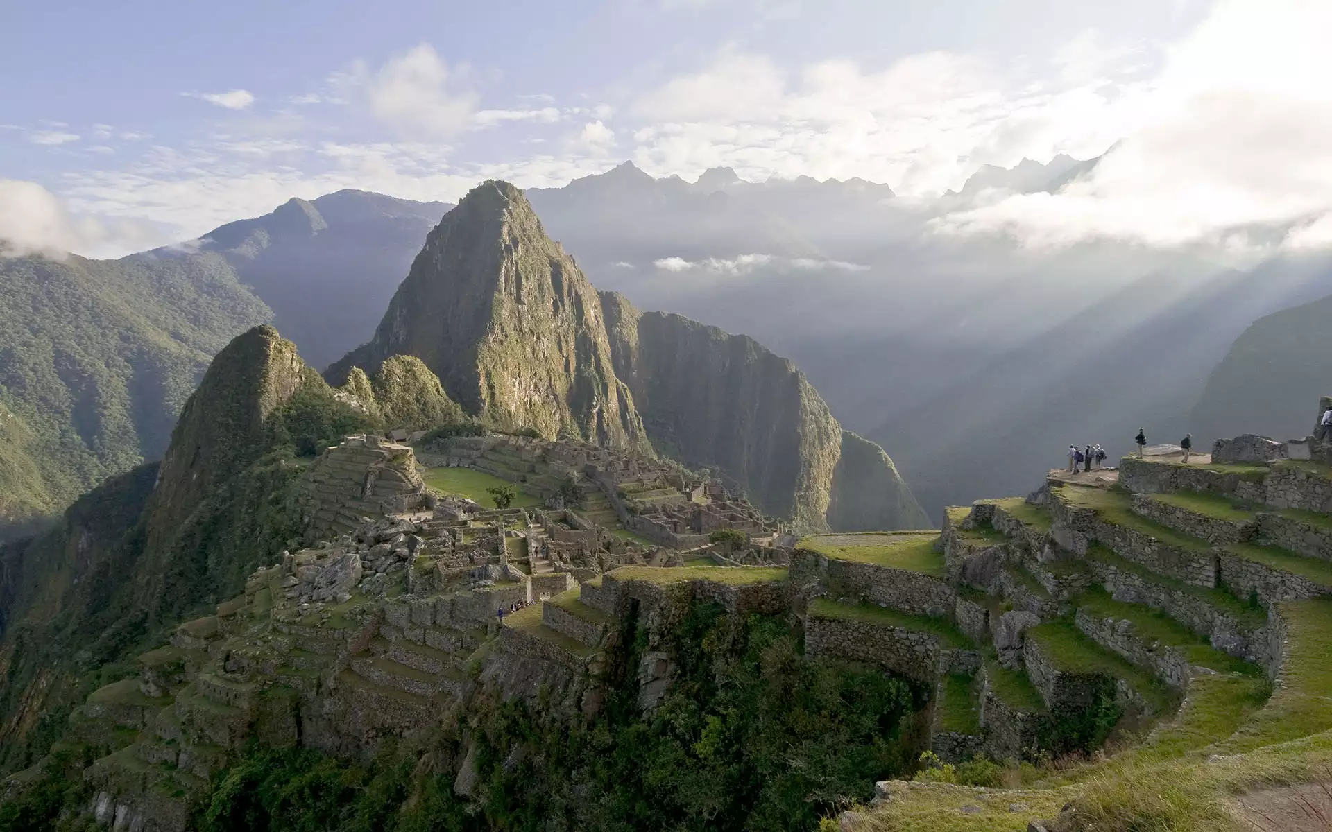 Sunbeams shine down on Machu Picchu and Huayna Picchu with a guided tour group seen standing on rock walls above the citadel