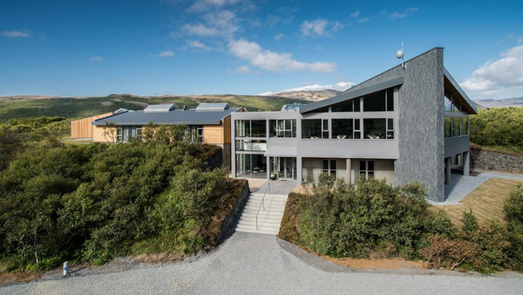 Aerial view of Hotel Husafell in Iceland with modern design of wood, cement, steel & large windows amid green hills & blue sky.