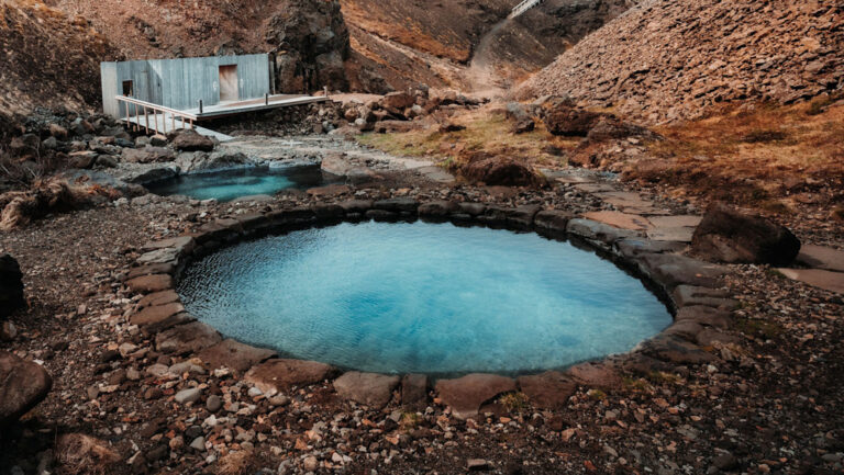 2 big natural hot spring pools with stone pathways & small wooden building by rust-colored hills at Hotel Husafell in Iceland.