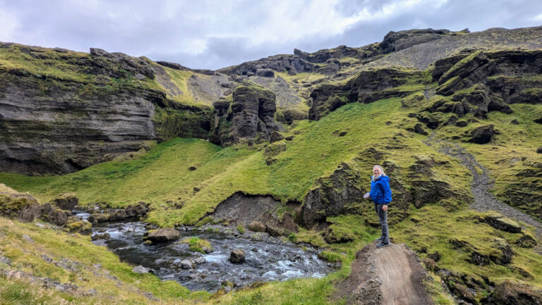 Woman in blue coat stands on overlook with bright green volcanic hills surrounding & river flowing below, in west Iceland.