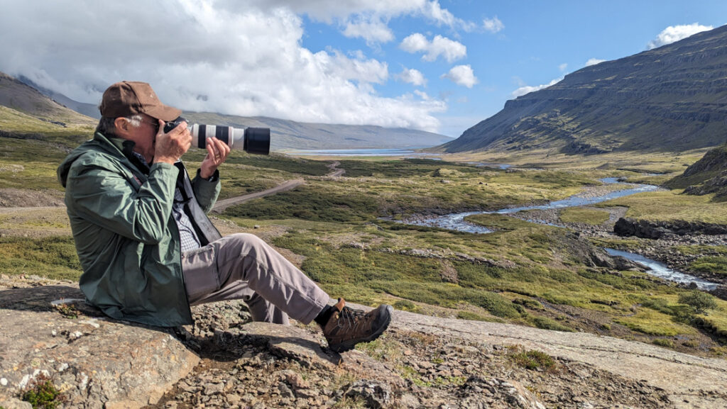 Man in green jacket holds large camera & photographs surrounding green hills & flowing rivers on a sunny day in west Iceland.