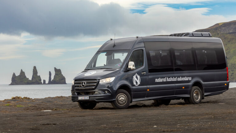 Gray Mercedes Sprinter van sits parked on beach with jagged islands offshore & green volcanic mountains behind, in Iceland.