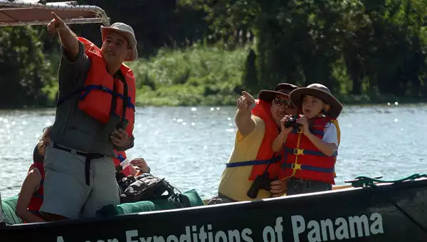 Father & guide point from a small green boat as boy looks on with binoculars in hand on sunny day of a tour of Panama.