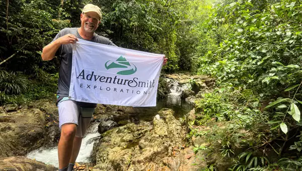 Man in shorts, t-shirt & hat holds a white flag with the AdventureSmith Explorations logo while standing beside a river with small waterfalls during a Panama tour.