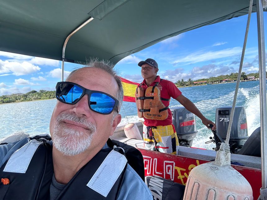 Man in blue mirrored sunglasses rides in boat driven by man in red shirt & swim trunks on a sunny day in Panama.