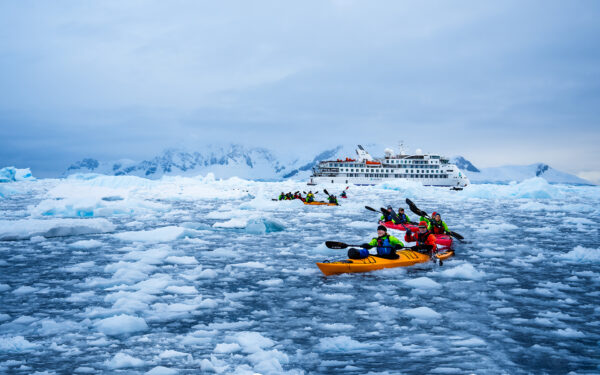 group of kayakers in brightly colored kayaks paddle through dense iceberg field. Greg Mortimer expedition ship anchored behind