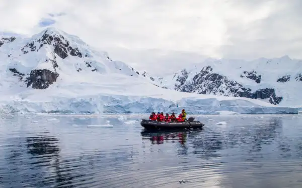 Group of Antarctica travelers Zodiac cruise in glassy water beside snowy peaks on a cloudy day.