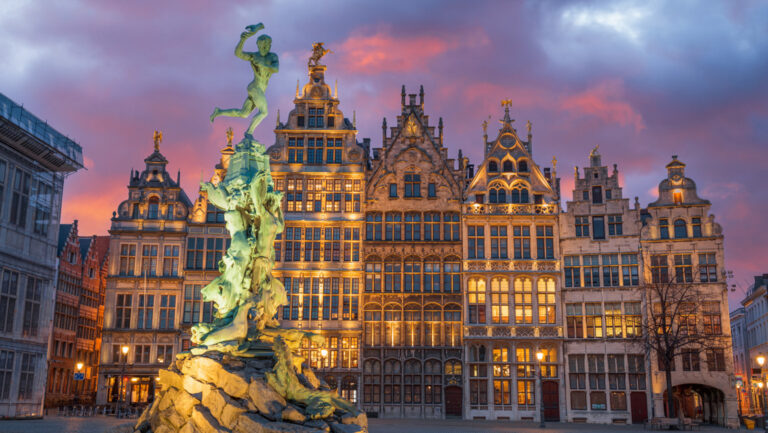 Green statue lit up in front of glowing gothic buildings under a pink & purple sky, seen on a Belgium river cruise.