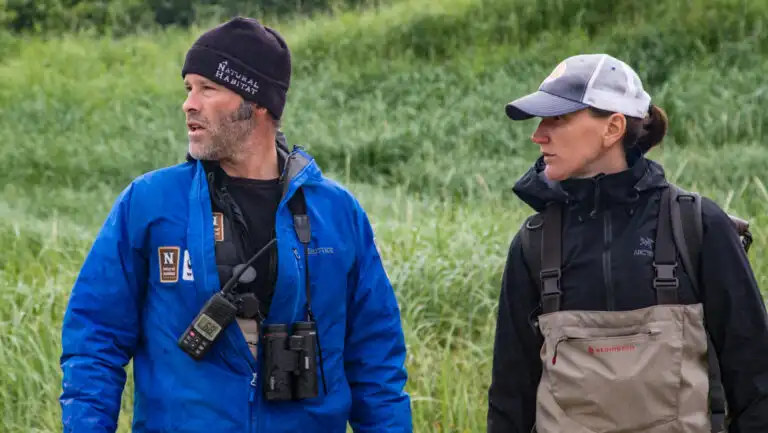 Male guide in blue jacket with radio stands beside woman in tan waders backed by grass on a tour for grizzly bear photography.