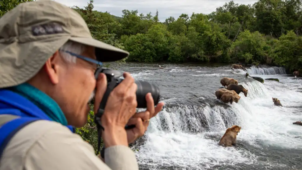 Man in tan hat & blue vest photographs grizzly bears fishing at river waterfalls on a Brooks Falls trip in Alaska.