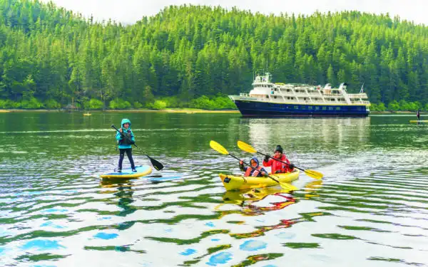 One kid on a paddleboard and a mom and son paddling in a yellow kayak in front of their small ship family cruise in Alaska