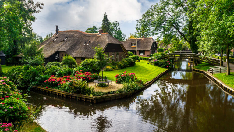 Glassy canal flows beside bright green grass, gardens of red flowers & shake-wood roofs on a sunny day of a Holland river cruise.