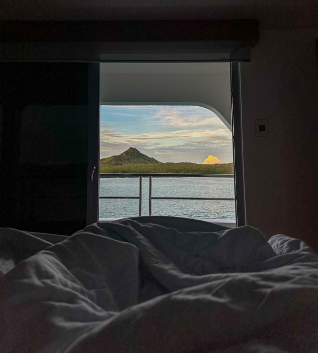 view from bed aboard small ship cruise looking out at the balcony views to the ocean and lush green landscapes