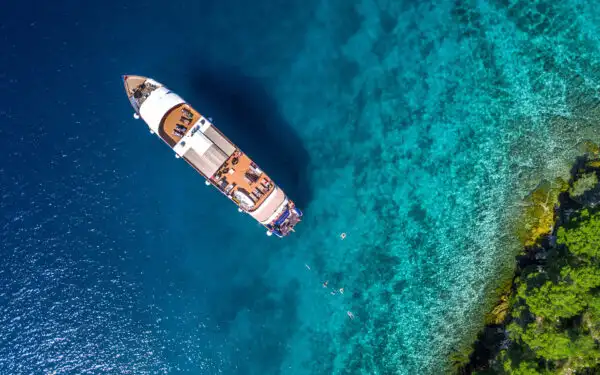 An aerial photo of a small ship anchored at a treed shoreline in the Mediterranean Sea with bright blue water and swimmers behind ship