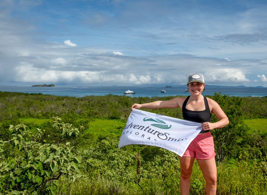 AdventureSmith Grace with pink hat and black shirt holding a flag standing at an overlook point in the Galapagos on a sunny day