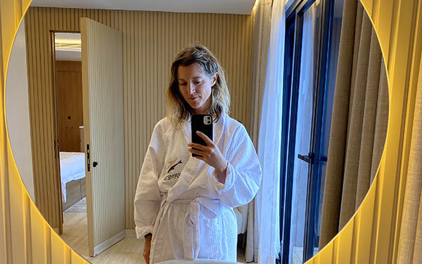 A female in a white robe takes a selfie in a light-lined circular mirror in a Hermes catamaran suite