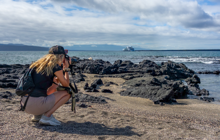 A female traveler squats to take an photo of iguanas with the Hermes catamaran in the water behind her