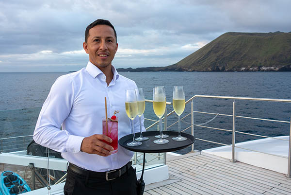 A bartender holds a tray of Champagne and an iced drink with a red cherry on the bow of a ship