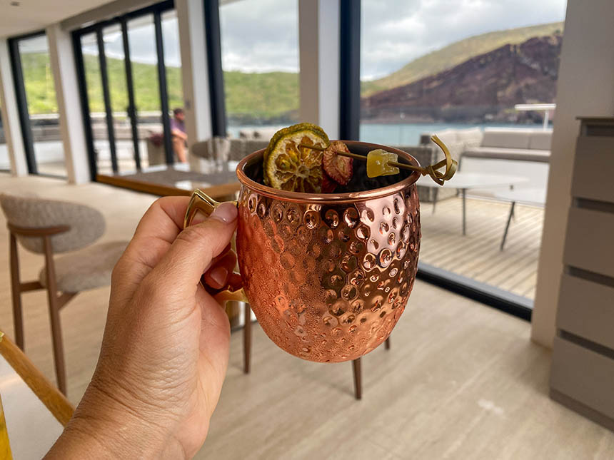 A hand holds out a moscow mule in a copper cup in the dining room of Hermes with a view out the stern deck
