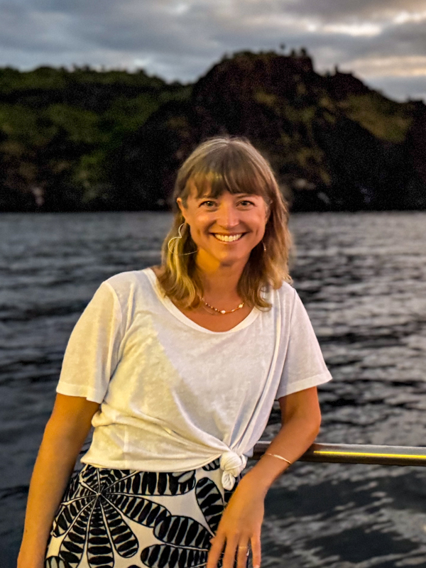 A female traveler in a white shirt leans on the railing of a ship in front of dark cliffs in the Galapagos