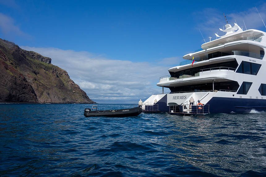 Hermes catamaran seen at anchor in the Galapagos near a rocky shore with a zodiac tied up to its stern